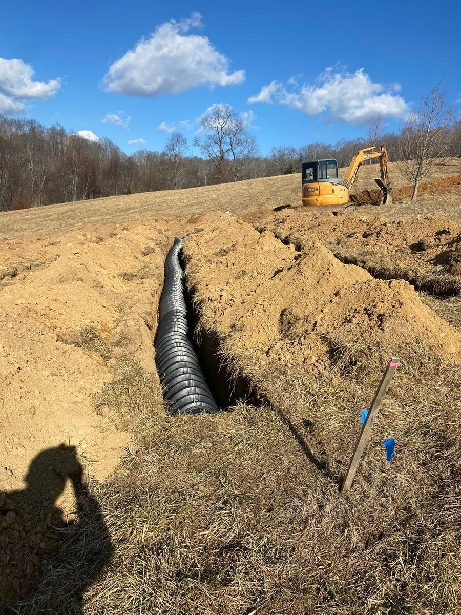 A culvert pipe being set into a freshly dug trench on a hillside with an excavator working behind it